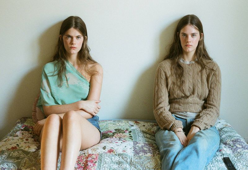 Two young women sit side-by-side on a floral quilt in an indoor setting, one in a turquoise top, the other in a tan sweater.
