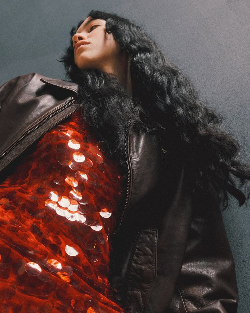 Stylish woman with long dark hair poses in a sequined dress and brown leather jacket against a textured grey-blue backdrop, shot from below.