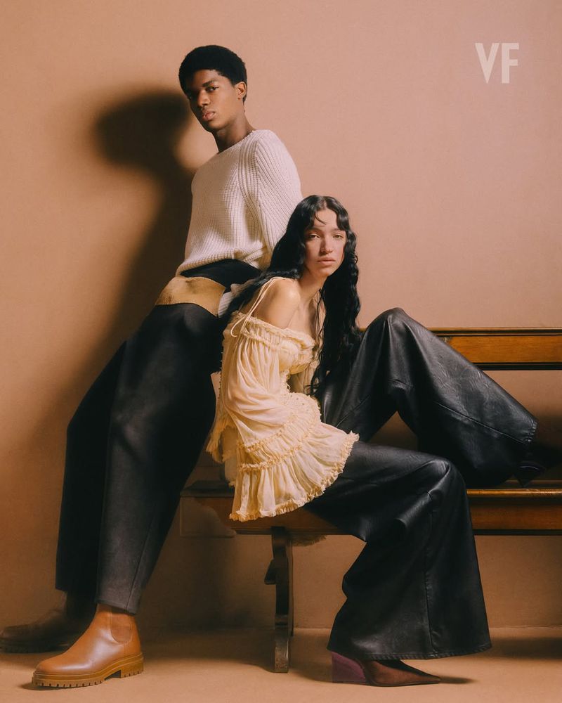 Two models posing together for Vanity Fair Italia, featuring a standing man in a white sweater and a seated woman on a wooden bench.