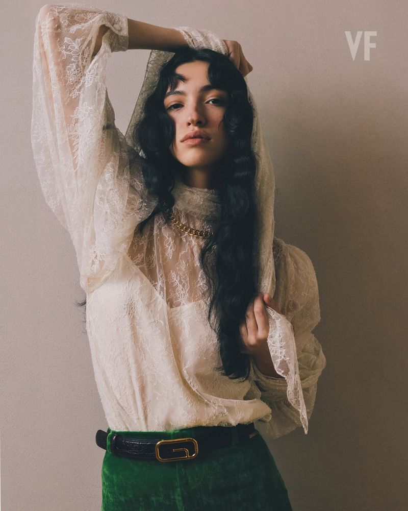 A woman with wavy dark hair poses indoors wearing a cream-colored lace top with long sleeves, green pants, and a gold chain necklace.