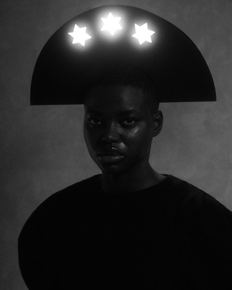 Portrait of a person with a black headdress featuring three glowing stars, set against a muted grey background in a studio.