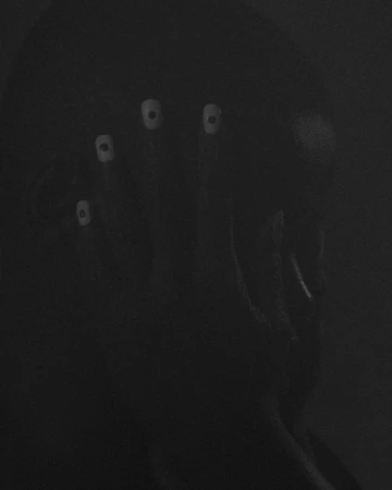 A person with pale, augmented eyes is seen in a dark environment, conveying an eerie and surreal visual, in a monochrome aesthetic.
