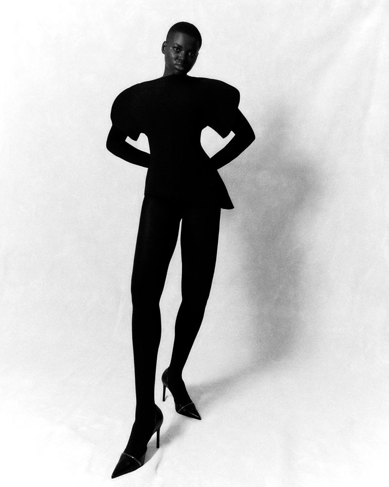 A stylish Black woman in a black outfit and heels strikes a pose against a white backdrop in a studio fashion photograph.