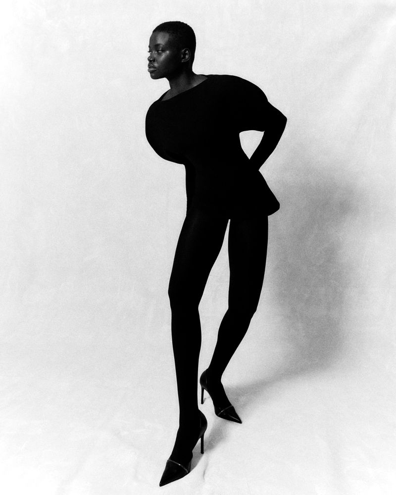 Black and white studio shot of a Black woman posing in a black dress, tights, and heels against a white backdrop.