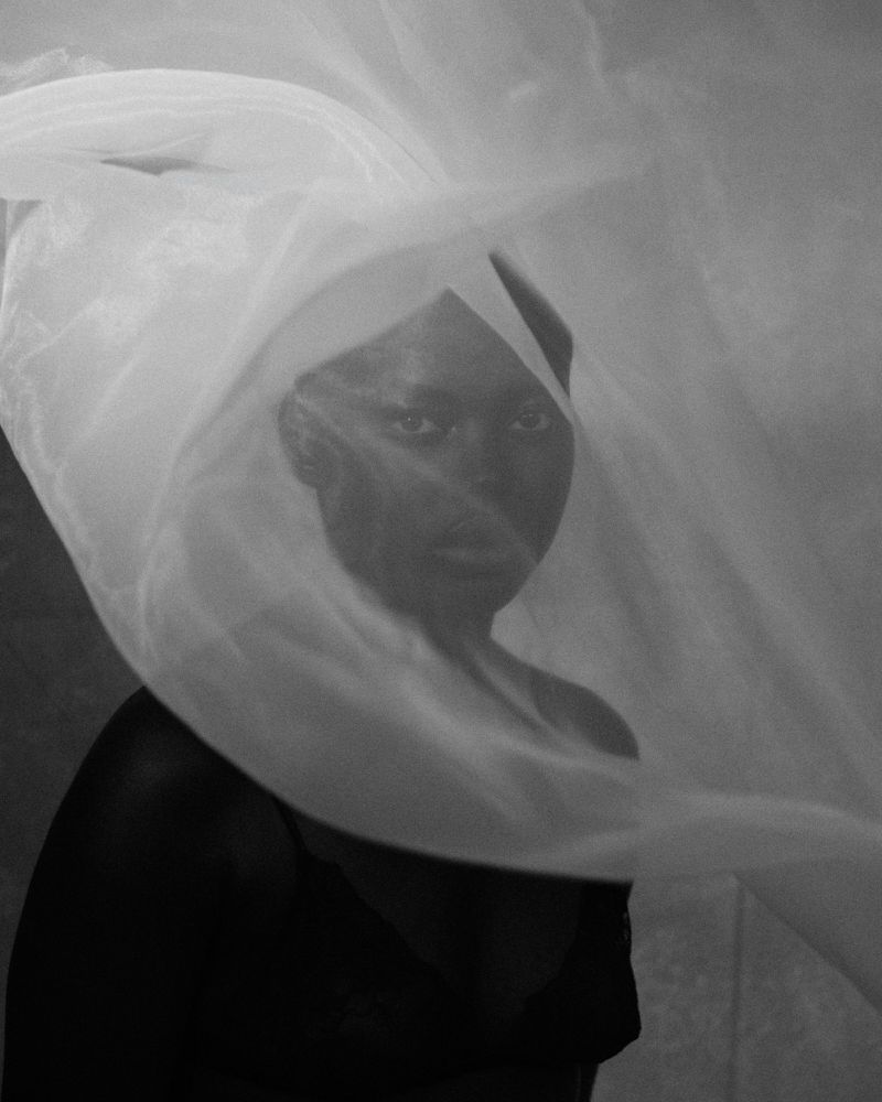 Monochrome portrait of a Black adult female with neutral expression, partially veiled in semi-transparent white fabric, medium shot.