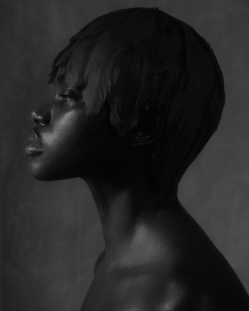 Striking monochrome portrait of a Black woman in profile, adorned with elegant feathers, set against a textured grey background.