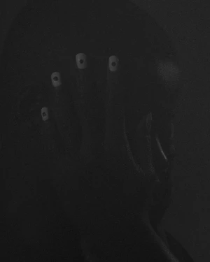 Close-up low-key portrait of a person in deep shadow with a hand raised to their face, displaying white nails with black dots.