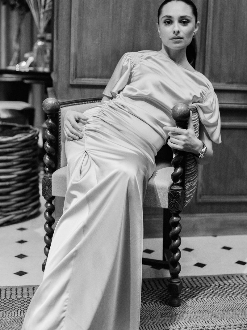 Monochrome portrait of a woman in a long dress seated on a chair in a neutral indoor setting, captured with natural lighting.