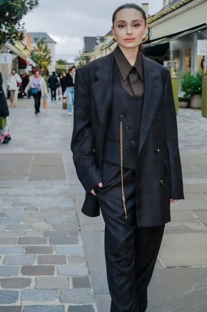 Woman in an oversized dark-grey suit with gold chain, standing on a cobblestone walkway in an outdoor shopping mall on an overcast day.