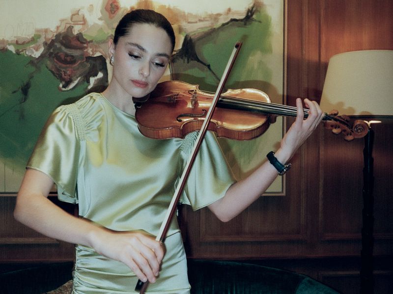 A woman in a light green dress plays a violin indoors, standing in front of an abstract painting and a lamp, creating an elegant, musical scene.