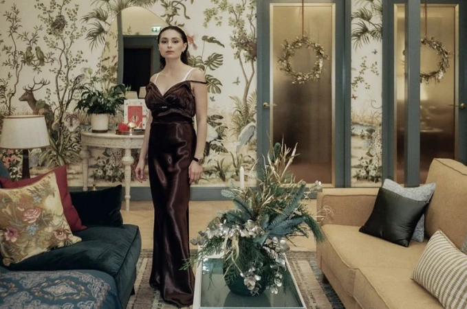 Elegant woman in a brown dress stands in an ornate interior with floral wallpaper, sofas, and decorative wreaths on glass doors.