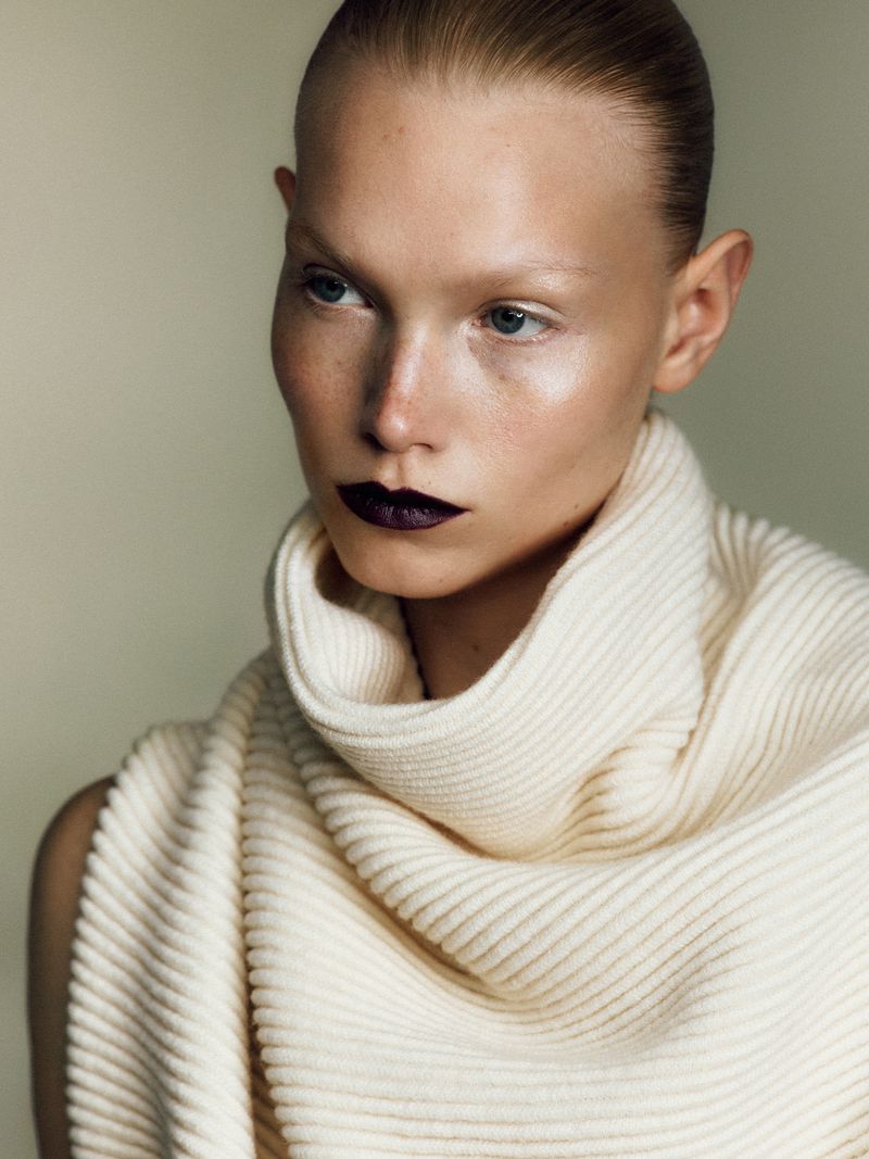 Close-up portrait of a fair-skinned adult female model with slicked-back hair, dark lipstick, wearing a textured white knit scarf indoors.