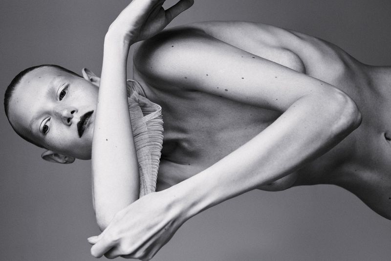 Monochrome fashion portrait of a model with dark lipstick and a pleated neckpiece posing against a neutral studio backdrop.
