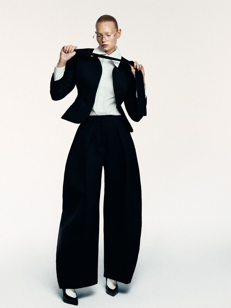 Woman poses in an oversized black suit with wide-leg pants, wearing a white shirt, black tie, white shoes, and gold glasses against white background.