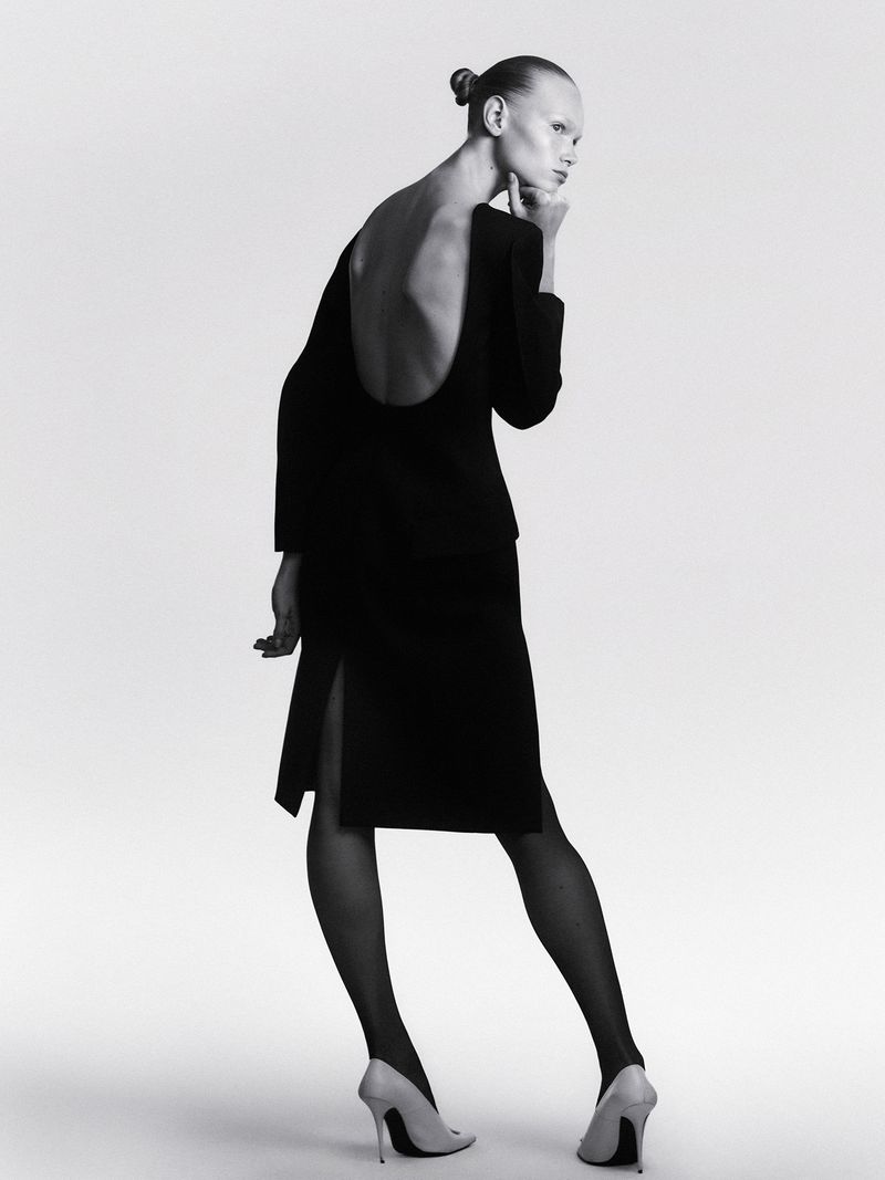 Woman in backless black dress and heels poses in a studio with minimal makeup, showcasing fashion photography and elegant style.