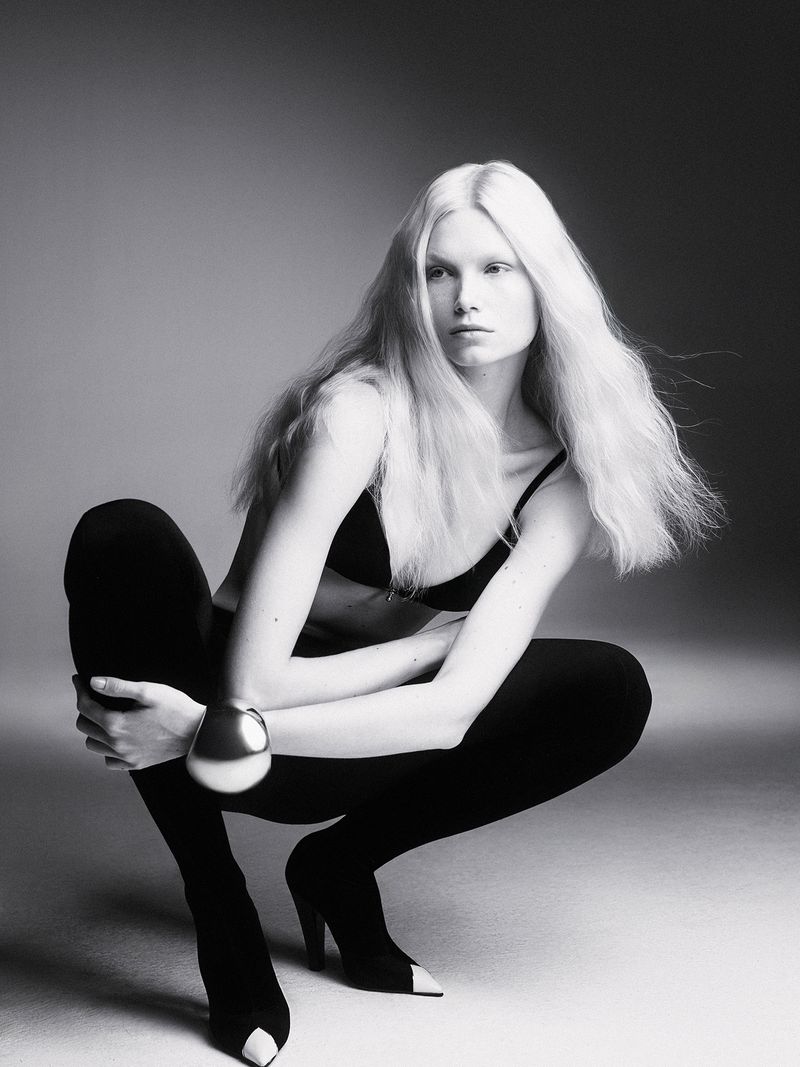 Monochrome studio portrait of a woman with long white hair, wearing a bra and leggings, kneeling in a crouching pose and looking forward.