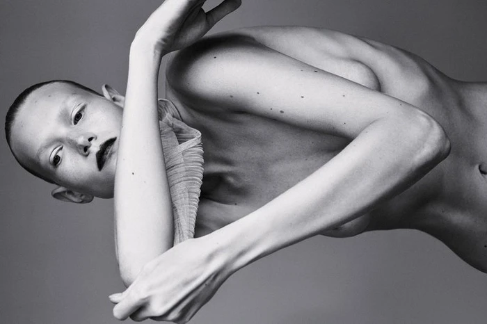 Monochrome fashion portrait of a model with dark lipstick and a pleated neckpiece posing against a neutral studio backdrop.
