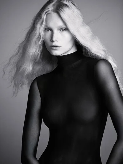 Black and white portrait of a blonde woman wearing a sheer black high-neck top against a smooth studio background.