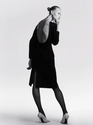 Model with slicked-back hair wearing a backless black midi dress, black tights, and white stiletto heels against a white backdrop.