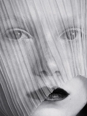 Black and white close-up portrait of a woman looking through a pleated sheer mesh veil with dark lipstick.