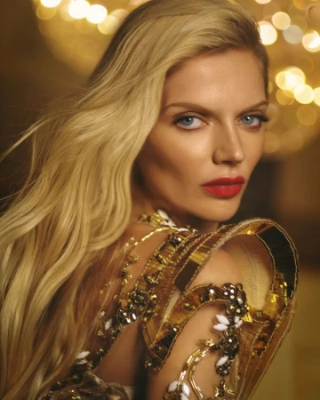 Blonde woman with red lipstick poses in golden, embellished gown against bokeh background. Close-up beauty shot.