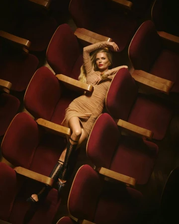 Blonde woman in a ruched nude dress reclining across rows of burgundy velvet auditorium chairs from an overhead angle.