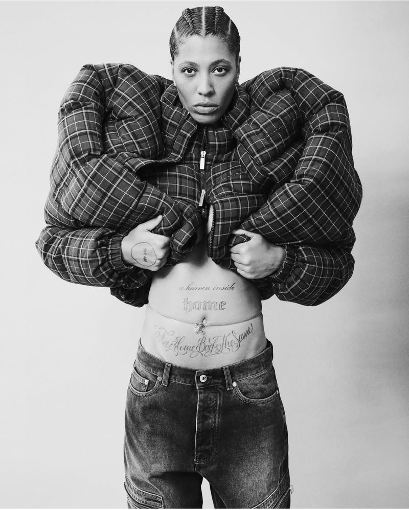 An adult female wearing a plaid jacket and jeans poses in front of a white background, exposing her torso with visible tattoos.