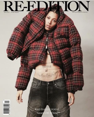 Model Kai Isaiah Jamal wearing an oversized red plaid puffer jacket and dark jeans against a neutral studio background.