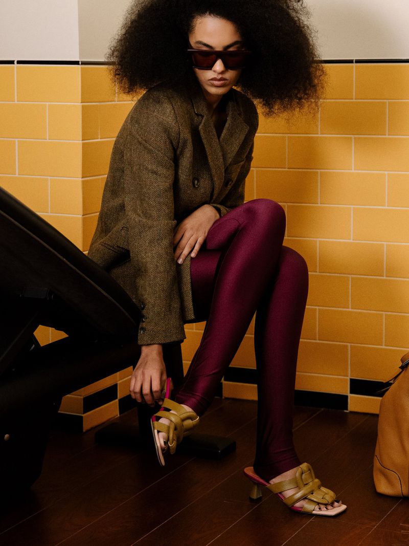 A woman with afro hair and sunglasses sits on a bench, wearing leggings and a jacket in front of yellow tiled wall, fastening sandals.
