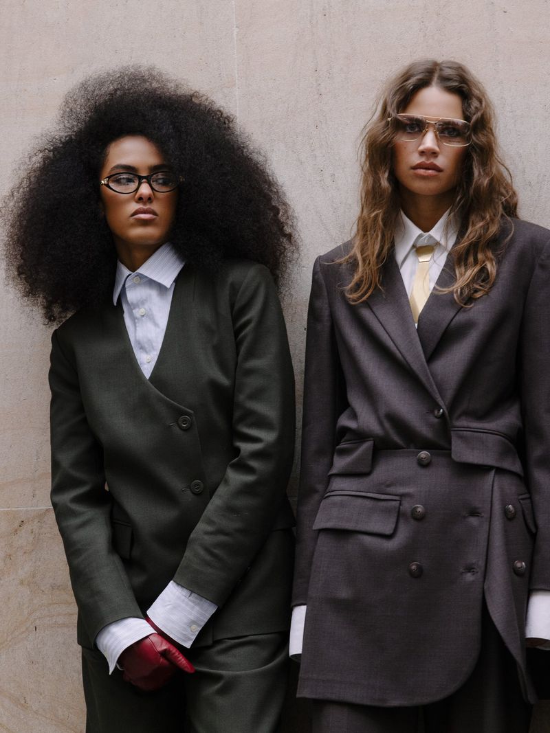 Two fashion models posing in tailored suits against a textured wall, showcasing contemporary style and design in a studio setting.