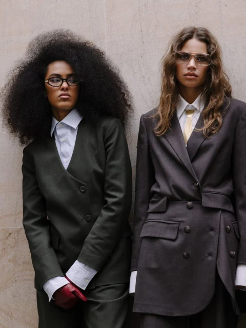 Two fashion models posing in tailored suits against a textured wall, showcasing contemporary style and design in a studio setting.