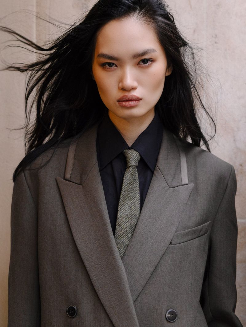 Portrait of a stylish Asian woman in a gray suit and black shirt, looking directly at the camera with a neutral expression.