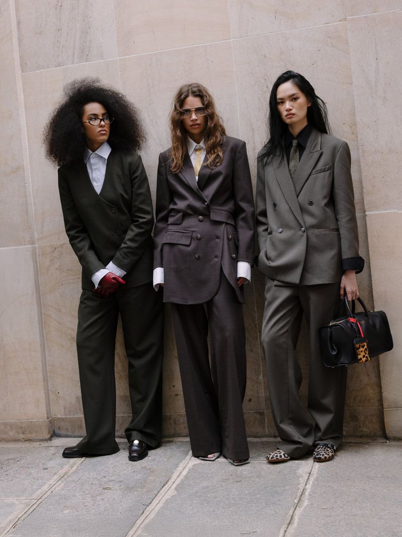 Three women stand in suits against a textured wall, showcasing tailored fashion with classic blazers and wide-leg pants in muted tones.