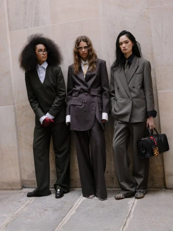 Three women stand in suits against a textured wall, showcasing tailored fashion with classic blazers and wide-leg pants in muted tones.