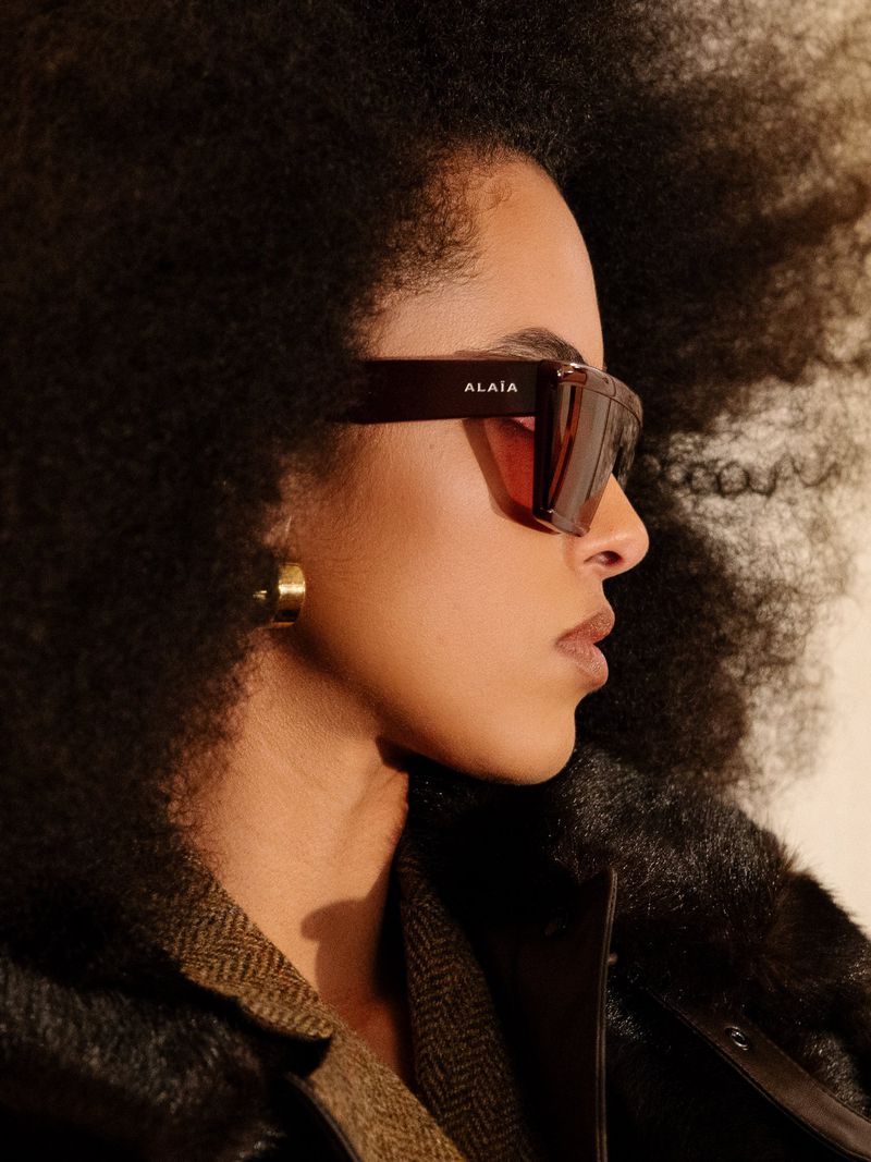 Profile of woman with large afro wearing sunglasses, gold hoop earring, and dark jacket against neutral background in a close up.