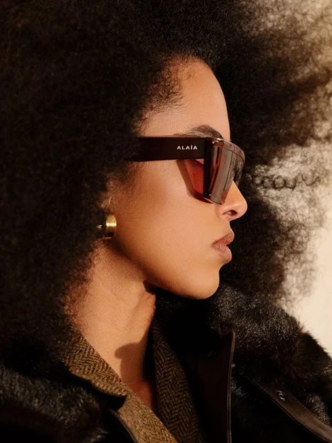 Profile of woman with large afro wearing sunglasses, gold hoop earring, and dark jacket against neutral background in a close up.