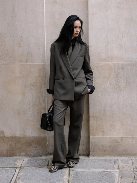 A woman poses against a textured wall wearing an olive pantsuit with a black purse and leopard print shoes in an outdoor setting.