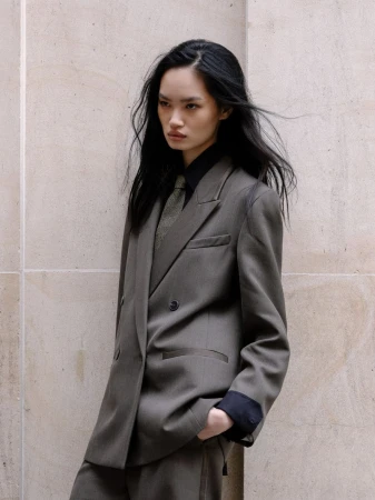 Model Nyouma Tacheboubet poses in an oversized taupe double-breasted suit and black shirt against a limestone wall.