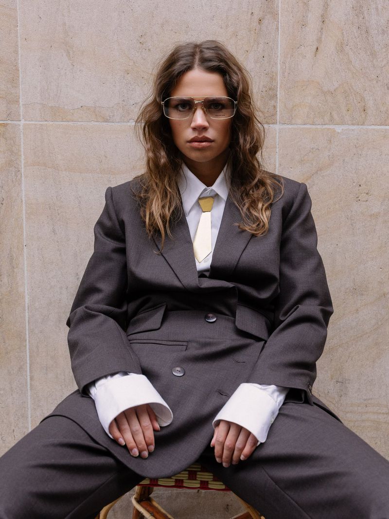A woman in a grey suit and glasses sits in front of a light tan textured wall in a fashion style image with a medium portrait framing.
