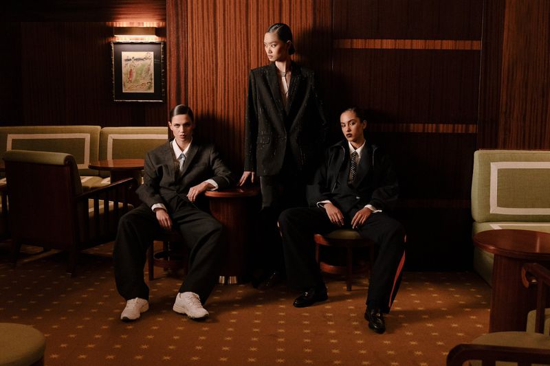 Three fashion models in suits pose indoors in a room with wood paneling and retro furniture, presenting a blend of classic and contemporary style.