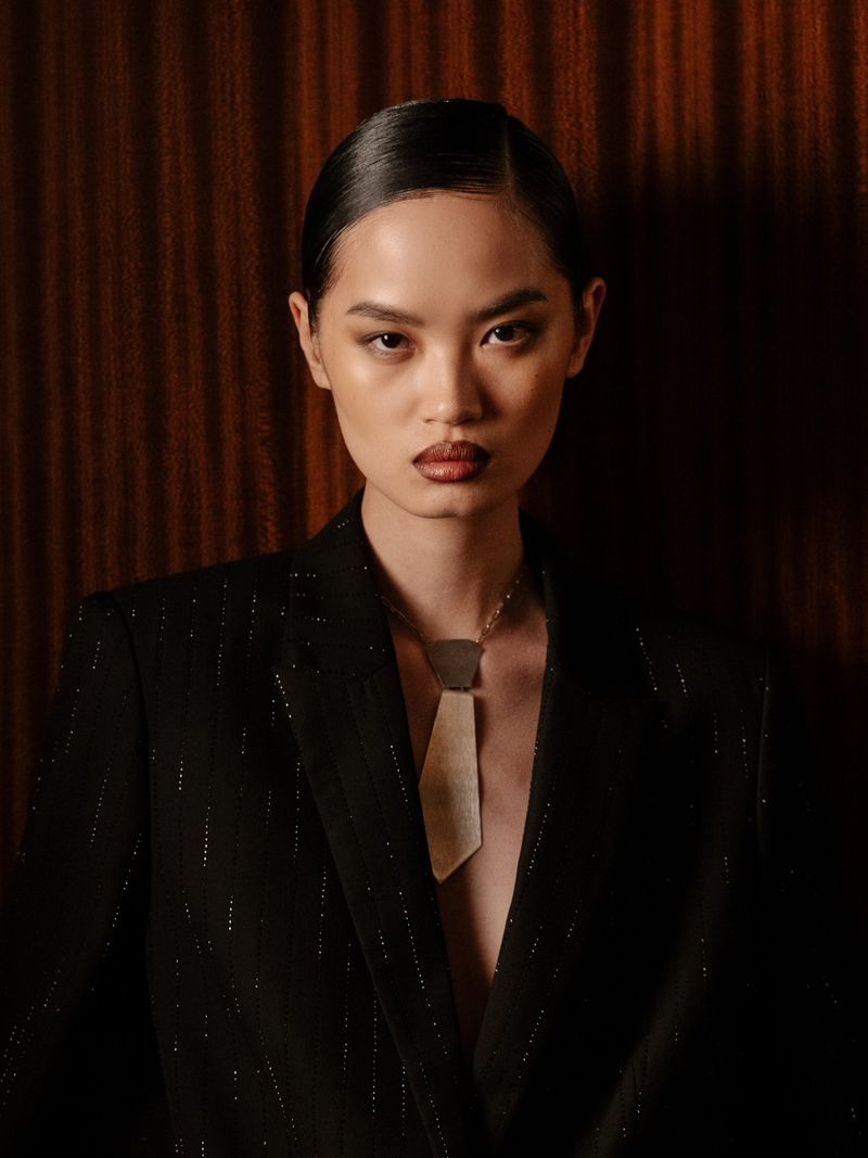 Close-up portrait of an Asian female fashion model wearing a black blazer and statement necklace, posing against a dark wood wall.