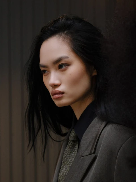 Portrait of an Asian woman with black hair wearing a dark green suit and patterned tie against a softly blurred, neutral background.