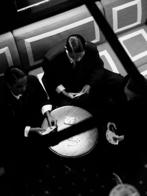 Three men play cards around a circular table indoors in a black and white photograph, captured from an overhead perspective.