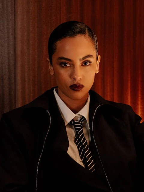 Stylish woman in a black suit with a striped tie looking at the camera with a dark lip in an indoor environment with neutral tones.
