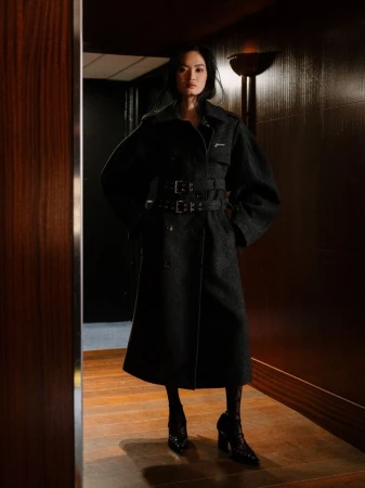 A woman in a dark belted coat stands indoors with a neutral expression. The lighting is soft and moody, accentuating the dark tones.