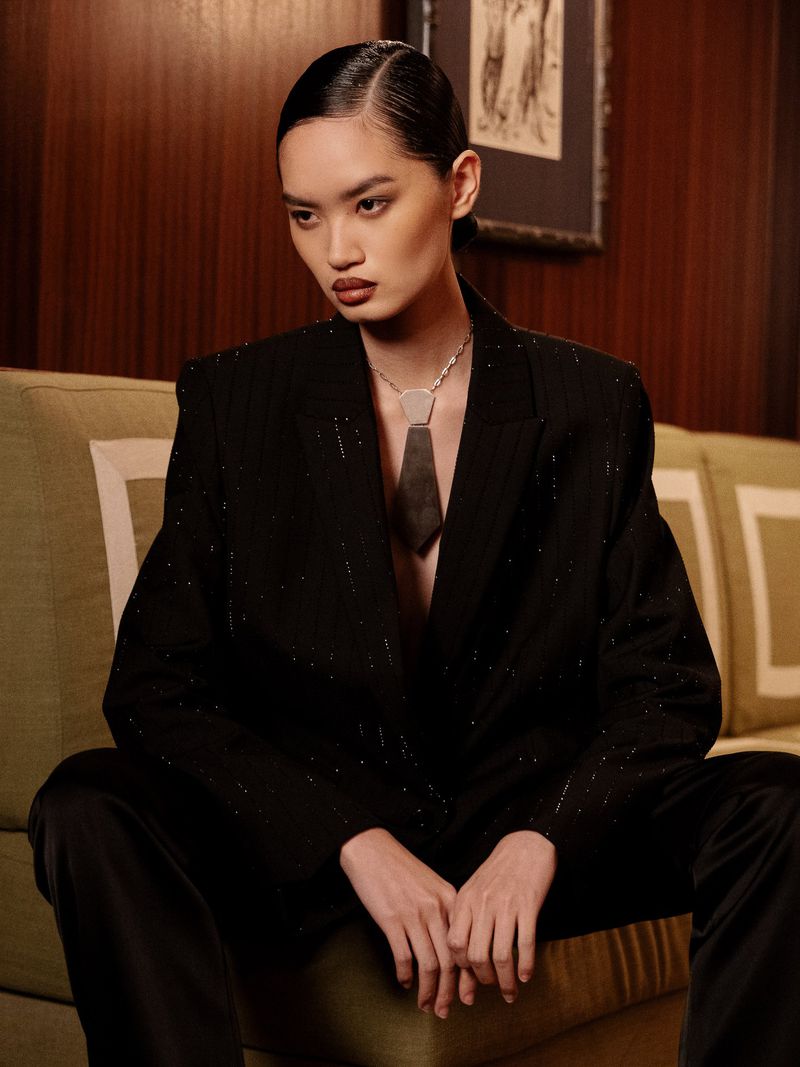 Elegant woman in black suit seated on a green sofa. She wears a statement necklace and poses in a refined indoor setting with wood wall.