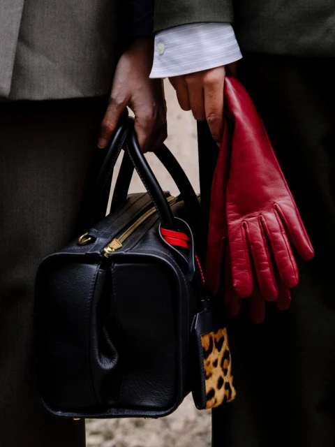 A person wearing a grey jacket is holding a black leather handbag, paired with bright red leather gloves, creating a bold contrast.