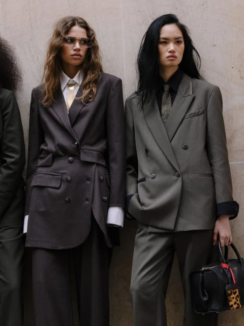 Two women in oversized blazers and ties stand indoors; one holds a black purse with a leopard-print tag in a high fashion pose.
