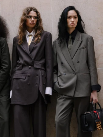 Two women in oversized blazers and ties stand indoors; one holds a black purse with a leopard-print tag in a high fashion pose.