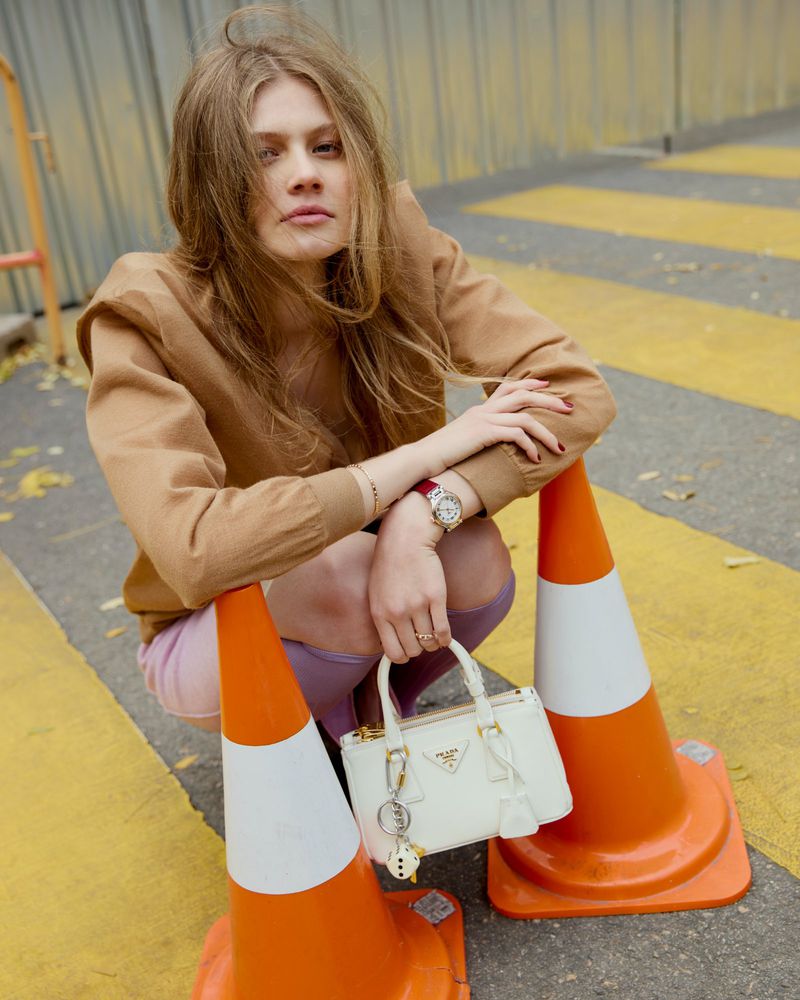 Young woman in casual outfit poses with Prada handbag between two orange traffic cones on a street with yellow road markings.